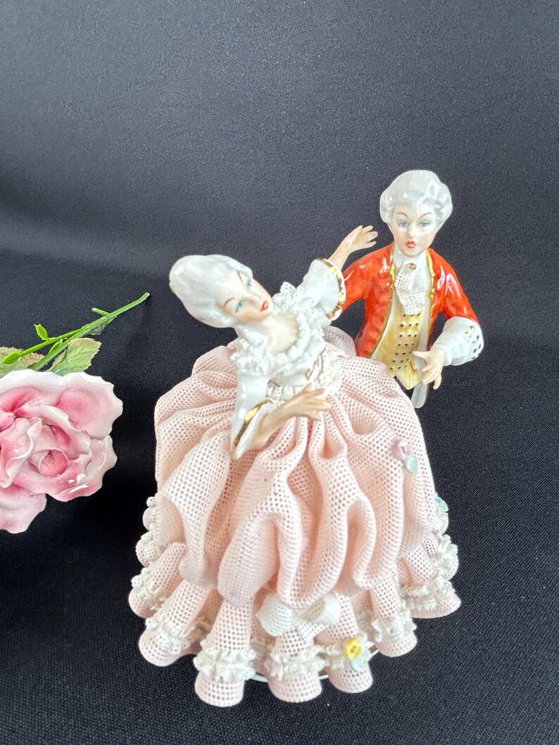 May include: A porcelain figurine set depicting a couple in 18th-century attire. The woman wears a pink dress with lace trim, while the man is in a red coat. The figures have detailed facial features and are posed as if dancing.