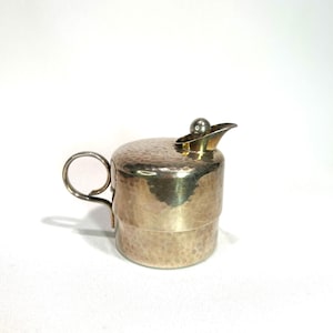 Könnte beinhalten: Eine kleine, silberfarbene Sahnekännchen mit gehämmerten Textur. Es hat einen Griff, einen Klappdeckel mit kleinem Ausguss und eine dekorative Kugel oben. Das Sahnekännchen ist zum Servieren von Flüssigkeiten bestimmt.