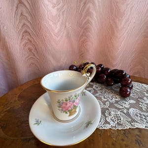 May include: A white porcelain teacup and saucer with a gold rim. The cup features a floral design with pink roses and a basket. The saucer has a small floral design.