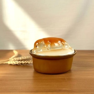 May include: A ceramic butter dish with a round, mustard-yellow base and a lid featuring a sculpted, glossy brown and white pig resting on top. The lid is white, and the pig figure has a smooth, shiny finish with detailed ears and snout.