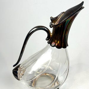 May include: A clear glass decanter with a silver-colored handle and spout. The decanter has a bulbous body with a swirl design and sits on a bronze-colored base. The spout has a decorative, flared design.