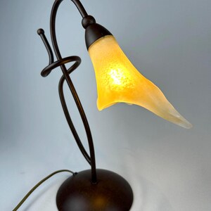 May include: A decorative table lamp with a bronze-colored metal base and stem. The lamp features a curved design with a yellow, tulip-shaped glass shade. The shade is lit from within, creating a warm glow. The lamp has a round base and a cord.