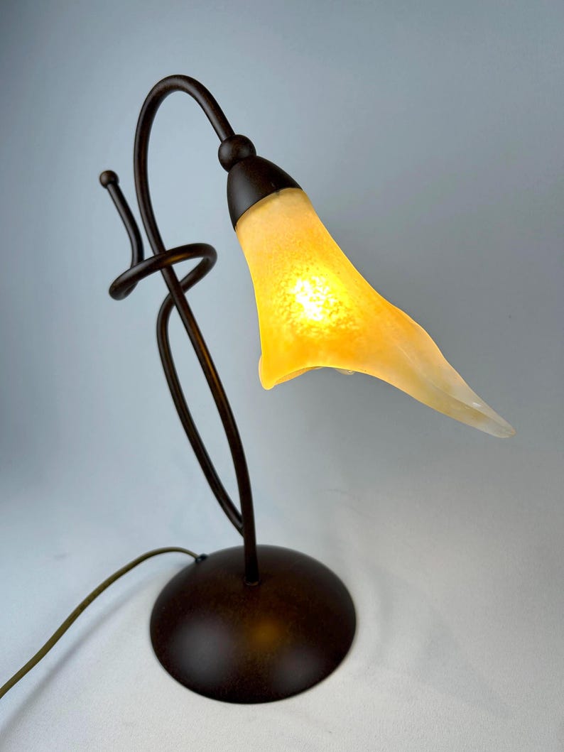 May include: A decorative table lamp with a bronze-colored metal base and stem. The lamp features a curved design with a yellow, tulip-shaped glass shade. The shade is lit from within, creating a warm glow. The lamp has a round base and a cord.