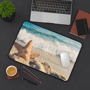 May include: A rectangular mouse pad featuring a beach scene with a starfish on the sand, waves, and blue water. A white computer mouse is on the pad. A laptop, notebooks, and a cup of coffee are also visible.