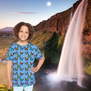 May include: A young person wearing a light blue t-shirt with a colorful butterfly pattern and light blue shorts. The background features a waterfall, mountains, and a bright sun in a clear sky.