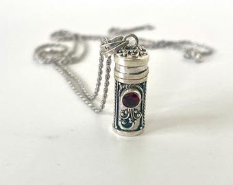 May include: A silver-colored pendant necklace featuring a cylindrical locket with ornate detailing. The locket has a red gemstone at the center and is suspended from a delicate chain. The design is intricate and the overall aesthetic is elegant.