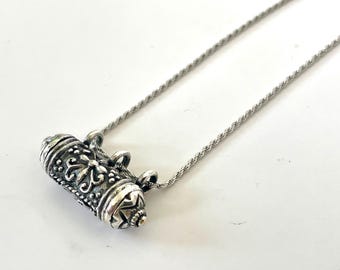 May include: A silver-colored pendant necklace. The pendant is cylindrical with ornate floral and dot patterns. The necklace chain is a thin, twisted rope design. The pendant is approximately 1.5 inches long.