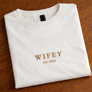 May include: White t-shirt with the word "WIFEY" embroidered in a gold color, along with "Est. 2026" below it. The shirt is folded neatly on a wooden surface. The t-shirt has a crew neck.