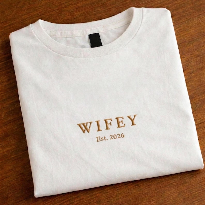 May include: White t-shirt with the word "WIFEY" embroidered in a gold color, along with "Est. 2026" below it. The shirt is folded neatly on a wooden surface. The t-shirt has a crew neck.