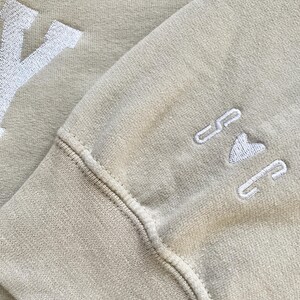 May include: A beige sweatshirt with white embroidered lettering that says "Y" on the left side and "I <3 NY" on the right side.