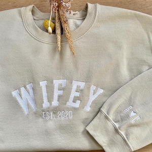 May include: A light beige sweatshirt with the word "WIFEY" embroidered in white, along with the text "EST. 2020". A small heart is embroidered on the sleeve.