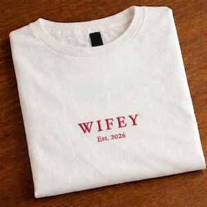 May include: A white t-shirt with the word "WIFEY" embroidered in red thread. Below the word is "Est. 2026". The shirt is folded neatly on a wooden surface.