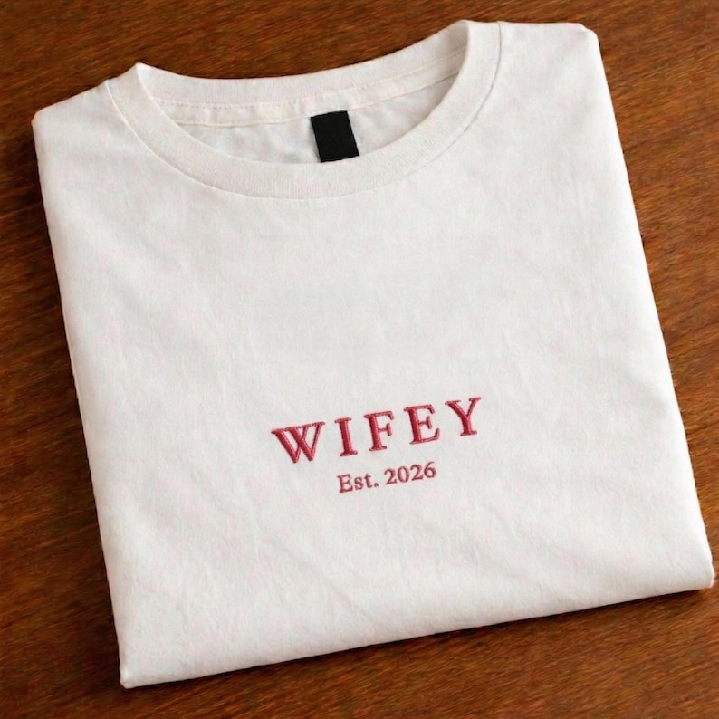 May include: A white t-shirt with the word "WIFEY" embroidered in red thread. Below the word is "Est. 2026". The shirt is folded neatly on a wooden surface.