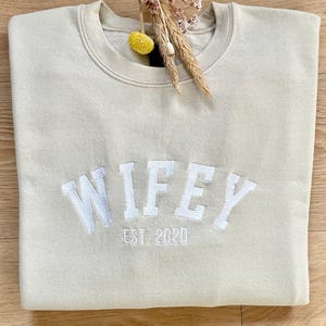 May include: A light beige sweatshirt with the word "WIFEY" embroidered in white with the text "EST. 2020" underneath.