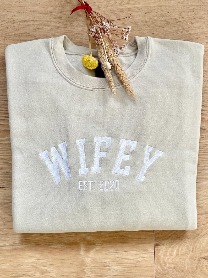 May include: A light beige sweatshirt with the word "WIFEY" embroidered in white with the text "EST. 2020" underneath.