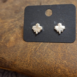 Puede incluir: Un par de pendientes de botón plateados con forma de hojas de arce. Los pendientes se muestran en una tarjeta negra con un solo agujero en la parte superior. La tarjeta descansa sobre una superficie de cuero marrón texturizado.