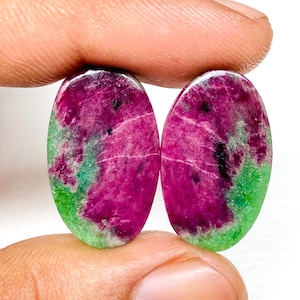 May include: Two oval-shaped gemstones with a vibrant color palette. The center of each stone is a deep magenta, transitioning to a bright green hue at the edges. The stones are held between fingers.