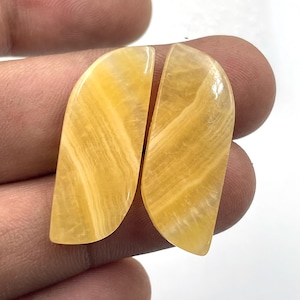Può includere: Due cabochon di pietre preziose lucidate, giallo dorato, con forma curva e allungata. Le pietre mostrano un motivo naturale e stratificato, e sono tenute in mano su sfondo bianco. I cabochon sono probabilmente per la creazione di gioielli.