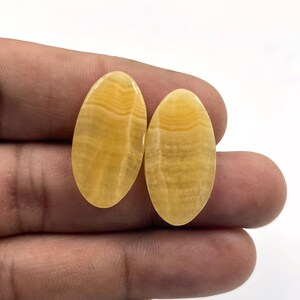 Può includere: Due pietre preziose ovali e lucide con una calda tonalità gialla. Le pietre mostrano un motivo naturale a strati, creando un effetto a righe. Le pietre preziose sono tenute in mano su uno sfondo bianco, evidenziando il loro colore e la loro consistenza.