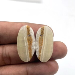 Può includere: Due pietre preziose ovali con base color crema e una fascia centrale beige e marrone. Le pietre sono tenute in mano su sfondo bianco. Le pietre hanno una superficie lucida e liscia.