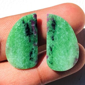 Könnte beinhalten: Zwei polierte Edelsteine, jeweils mit abgerundeter Form und leuchtend grüner Farbe. Die Steine weisen dunkle schwarze und tiefrote Einschlüsse auf, die einen auffälligen Kontrast erzeugen. Die Edelsteine werden in einer Hand gehalten und zeigen ihre natürliche Schönheit.