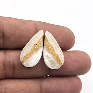 Può includere: Due pietre preziose a forma di goccia. Ogni pietra ha una base bianca con una fascia centrale di materiale granulare color oro. Le pietre sono tenute in mano su uno sfondo bianco. Queste pietre preziose potrebbero essere utilizzate per la creazione di gioielli.