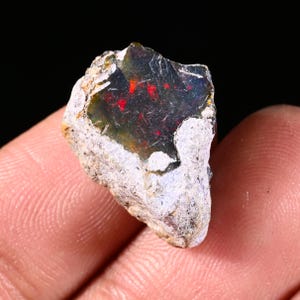 Op de afbeelding: Een ruwe, ongepolijste opaal edelsteen met een donker, doorschijnend lichaam met levendige rode en oranje kleurflitsen. De steen heeft een ruwe, getextureerde, lichtgrijze buitenmatrix die het gepolijste, donkere, iriserende middengedeelte omringt.