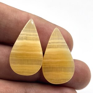 Può includere: Due pietre preziose lucide a forma di goccia. Le pietre sono di un colore giallo caldo con bande orizzontali, creando un effetto a righe. Le gemme sono tenute in una mano, mostrando le loro superfici lisce e riflettenti. Adatto per la creazione di gioielli.