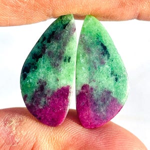 Op de afbeelding: Twee gepolijste edelstenen, elk met een druppelvorm. De stenen vertonen een mix van kleuren, waaronder een levendig magenta aan de basis, overgaand in groen en zwart naar de top. De edelstenen worden tussen vingers gehouden.
