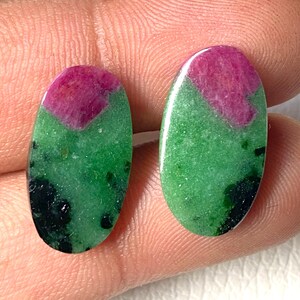 May include: Two oval-shaped gemstones. Each stone features a vibrant magenta section at the top, transitioning to a rich green base with dark flecks. The stones are smooth and polished, showcasing natural color variations.