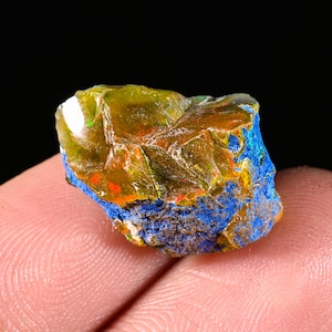 Puede incluir: Un espécimen mineral en bruto que presenta una sección superior translúcida de color ámbar con destellos internos de luz roja y verde, sobre una base rugosa de color azul profundo con una superficie granular y texturizada, mostrando formaciones geológicas.