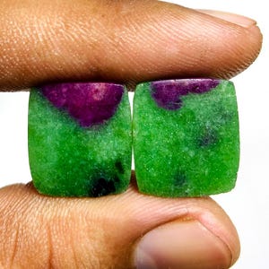 May include: Two square-cut gemstones with rounded corners. The stones have a vibrant green base with a deep purple patch at the top. The gemstones are held between fingers, showcasing their polished surfaces and natural colour variations.