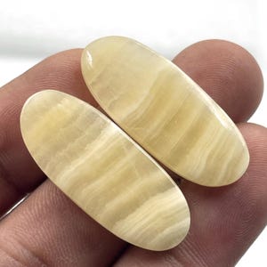 Può includere: Due pietre preziose ovali e lucide, di colore giallo crema, con sottili bande parallele. Le pietre sono tenute in mano, evidenziando le loro superfici lisce e riflettenti. Queste pietre preziose sono probabilmente utilizzate per gioielli o scopi decorativi.