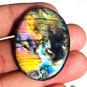 Può includere: Pietra preziosa di labradorite di forma ovale con superficie lucida. La pietra mostra un vibrante gioco di colori, tra cui tonalità gialle, rosa, blu e verdi. La gemma è tenuta in mano, mostrando le sue qualità iridescenti.