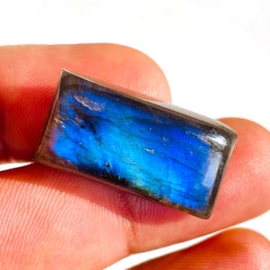 May include: A rectangular labradorite gemstone with a polished surface. The stone displays a vibrant play of color, with shades of blue and hints of green. The gemstone is held between fingers, showcasing its natural beauty.