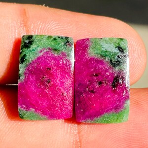 May include: Two rectangular ruby zoisite gemstones featuring vibrant magenta centers surrounded by mottled green and black mineral inclusions. The stones have a smooth, polished surface with a flat, rectangular shape and distinct color contrast.