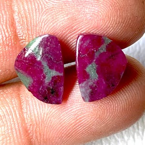 Puede incluir: Dos gemas de rubí pulidas, de color rojo intenso con inclusiones verdes. Las gemas se sostienen entre los dedos, resaltando su textura natural. Ideales para la creación de joyas.