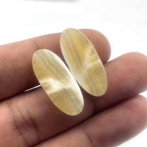 Può includere: Due pietre preziose ovali con una tonalità giallo crema traslucida e venature bianche. Le pietre sono tenute in una mano, mostrando la loro superficie lucida e i loro motivi naturali. Le pietre preziose sono probabilmente utilizzate per gioielli o scopi decorativi.