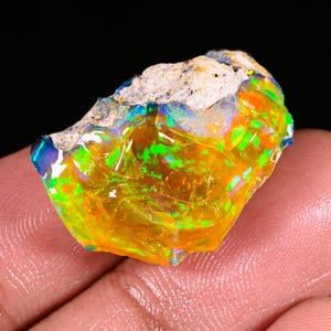 May include: A raw, unpolished opal gemstone featuring a vibrant play-of-color with intense flashes of neon green, bright orange, and deep blue. The stone has a rough, textured, light-colored matrix top edge and a smooth, translucent, glossy surface.