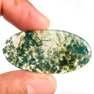 May include: An oval moss agate gemstone. The stone has a translucent base with intricate patterns of green, white, and yellow inclusions, resembling a miniature landscape. The gemstone is held between two fingers.