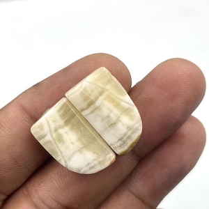 Può includere: Due pietre preziose lucide, semicircolari, con un motivo a bande bianco crema e marrone chiaro. Le pietre sono tenute in una mano su sfondo bianco. Le pietre hanno una finitura liscia e lucida.