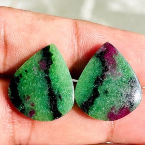 Peut inclure: Deux pierres précieuses en forme de goutte d'eau, avec une base verte vibrante, des veines noires et des éclats rouge foncé. Les pierres sont tenues dans une main, mettant en valeur leurs surfaces polies et leurs motifs naturels. Idéal pour la joaillerie.