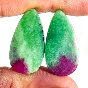 Może przedstawiać: Dwa kamienie szlachetne w kształcie łezki. Każdy kamień ma żywy zielony odcień u góry, przechodzący w głęboką fuksję u podstawy. Kamienie mają wypolerowaną powierzchnię z subtelnymi zmianami koloru i faktury.