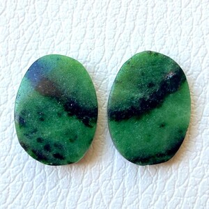 Può includere: Due pietre preziose verdi di forma ovale con inclusioni nere e marroni scure. Le pietre hanno una superficie lucida e sono poste su uno sfondo bianco. Adatto per la creazione di gioielli.