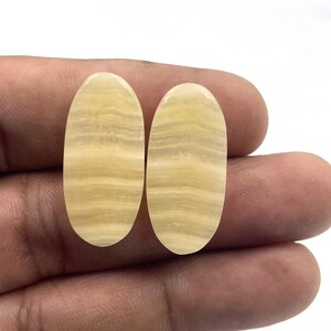 Può includere: Due pietre preziose ovali e lucide, di colore giallo crema e con motivi a bande. Le pietre sono tenute in una mano, mettendo in risalto le loro superfici lisce e riflettenti. Queste pietre sono adatte per la creazione di gioielli.