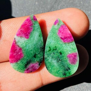 Può includere: Due pietre preziose a forma di goccia. Le pietre presentano una sorprendente combinazione di rosa brillante e verde intenso, con un motivo maculato. Le pietre preziose sono lucide e lisce, mostrando la loro bellezza naturale.