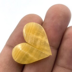 Può includere: Due pietre preziose a forma di cuore, lucidate, nei toni del giallo e dell'oro, sono tenute in una mano. Le pietre hanno una finitura liscia e lucida e un motivo naturale venato. La forma a cuore è formata da due pezzi separati.