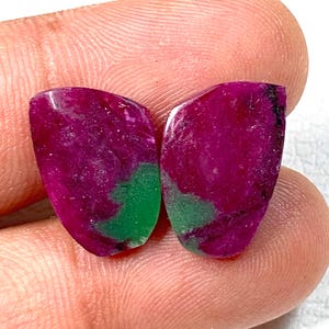 Op de afbeelding: Twee gepolijste edelstenen met een diepe magenta tint, geaccentueerd door vlekken levendig groen. De stenen hebben een glad, reflecterend oppervlak en een onregelmatige vorm. De edelstenen worden tegen een neutrale achtergrond gepresenteerd.