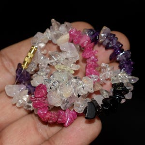 Puede incluir: Una pulsera de múltiples hilos compuesta por fragmentos irregulares de piedras preciosas pulidas en varios colores, incluyendo transparente, rosa, magenta intenso, morado y negro. La pulsera cuenta con un cierre de rosca cilíndrico dorado.