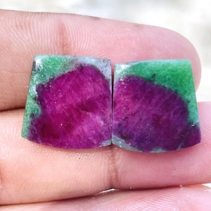 Puede incluir: Dos piedras preciosas de zoisita trapezoidales que presentan un centro de rubí magenta vibrante rodeado por una matriz mineral de color verde brillante. Las piedras tienen una superficie lisa y pulida con límites de color distintos entre las secciones rosa oscuro y verde.